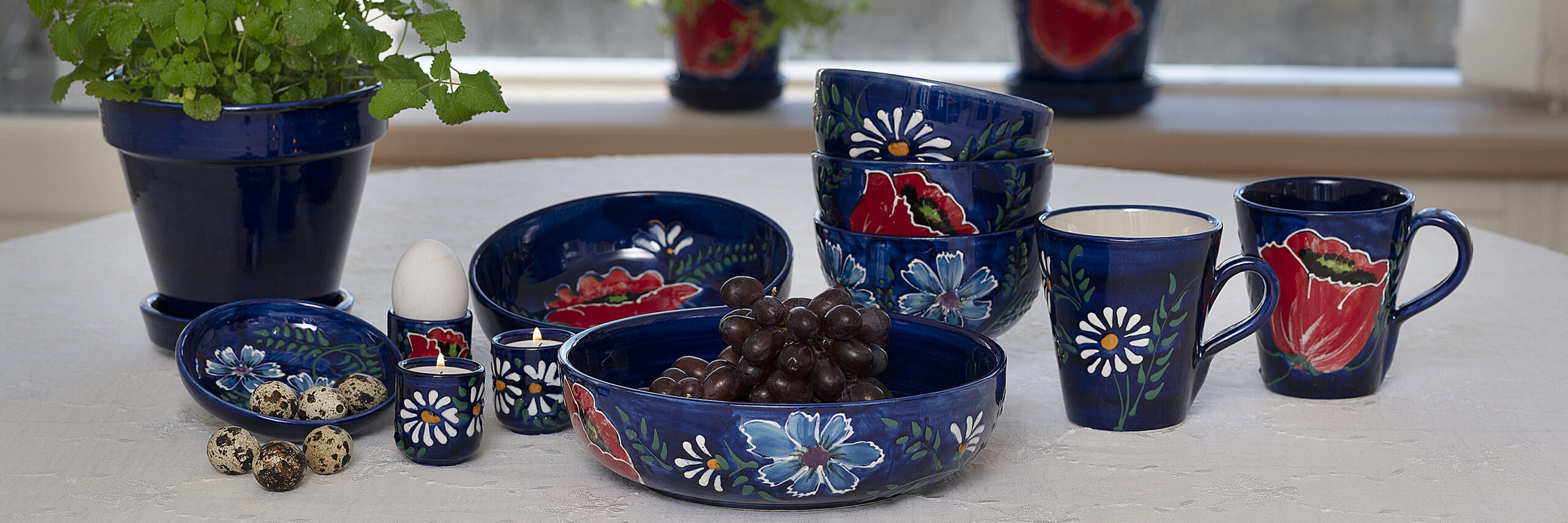 Casa Jada Keramik Spansk Forsidebilledet med Anna-serien. Blå bundfarve med valmuer, margeritter og kornblomster på hvid dug. Urtepotter i baggrund.
